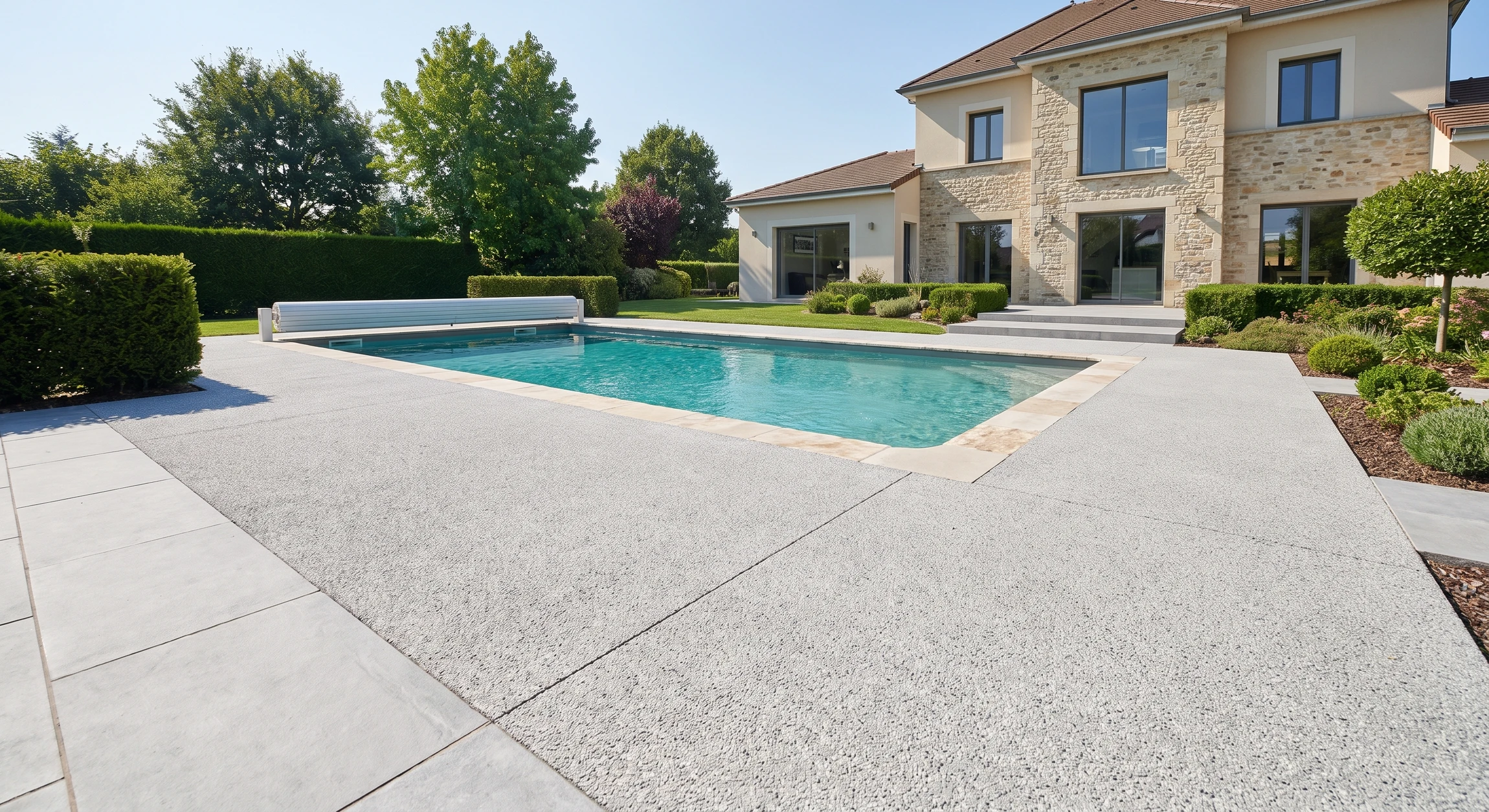 Photographie réelle en grand format montrant une réalisation d'aménagement extérieur complète d'ENT Prévitali : une terrasse pavée en béton désactivé ou dallage gris clair, adjacent à une piscine enterrée avec revêtement turquoise, margelles en pierre, et un volet roulant automatique. Au premier plan, des pavés ou dalles de qualité en pose jointive. En arrière-plan, une maison résidentielle contemporaine, végétation d'encadrement (arbres, haies taillées), ciel dégagé. La composition doit mettre en avant l'excellence du travail : surfaces planes parfaites, joints nets, intégration harmonieuse piscine-terrasse-maison, finitions soignées. Aucun personnage, aucun texte.